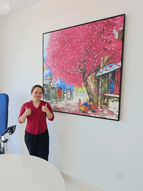 A woman giving a thumbs-up next to a large painting of a pink flowering tree and a street scene.