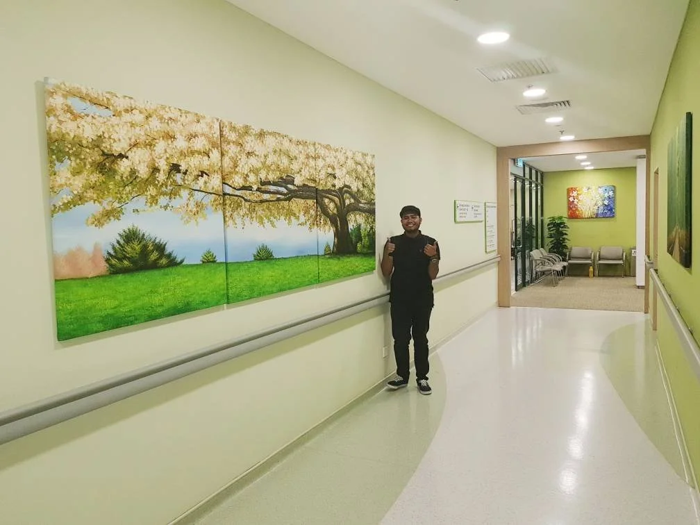 Person standing in a hallway near a landscape painting of a tree with yellow leaves, green grass, and a blue sky, inside a building with light green walls and a seating area with chairs and a colorful painting on the wall.