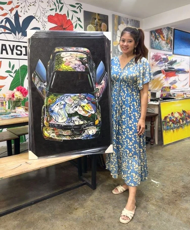 A woman in a blue floral dress standing next to a painting of a car, which features colorful, cartoon-style faces on its surface. The room is decorated with various paintings and art supplies.