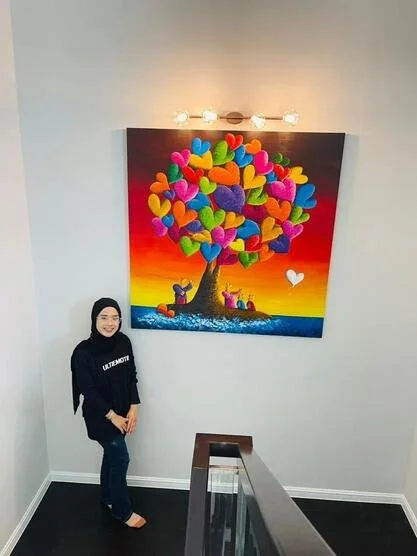 Woman standing in front of a colorful painting of a tree with heart-shaped leaves on a sunset background, hanging on a plain white wall.