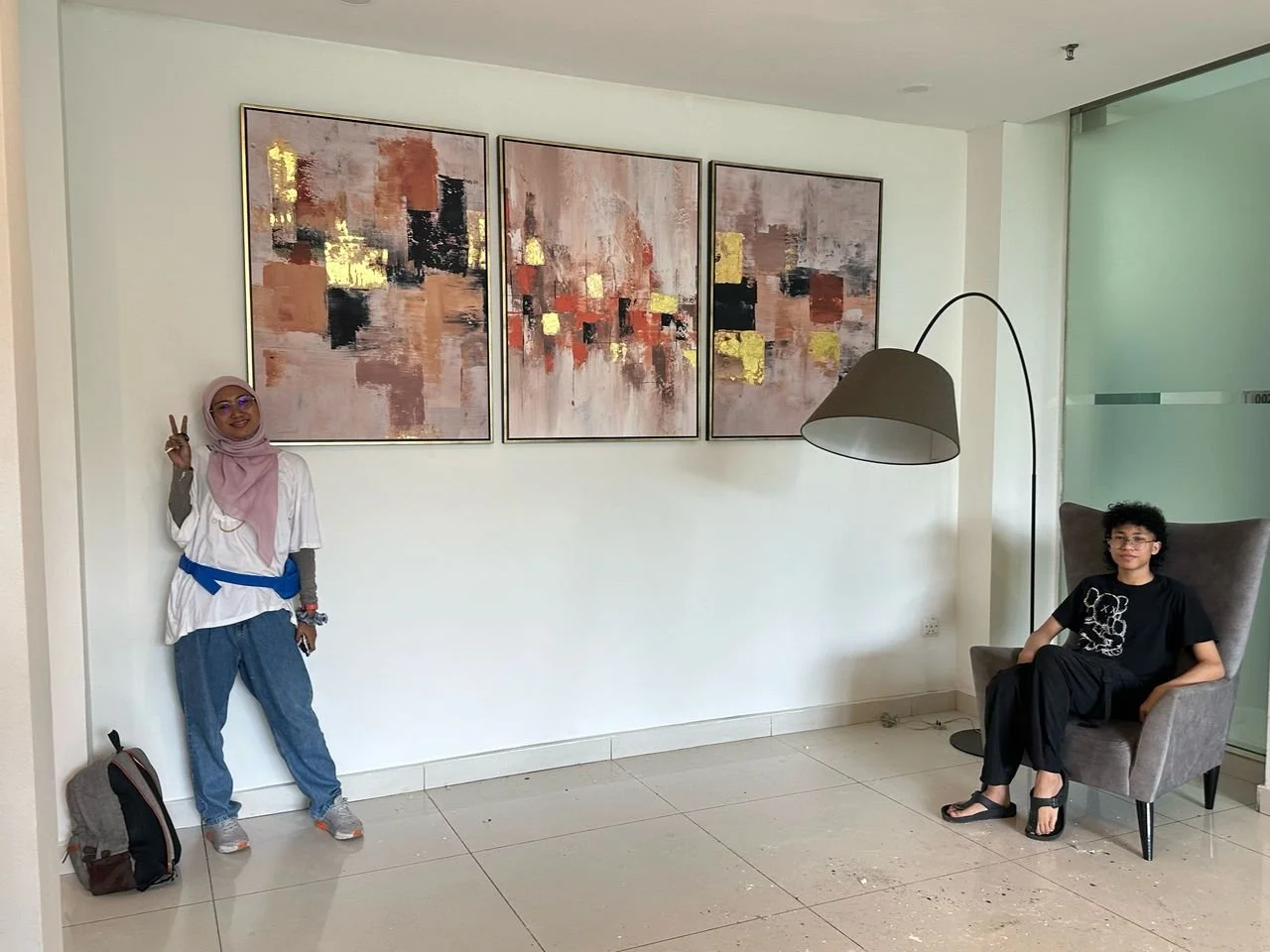 A woman wearing a pink hijab and white top making a peace sign standing next to a man sitting in a gray armchair, with colorful abstract paintings on the wall behind them.