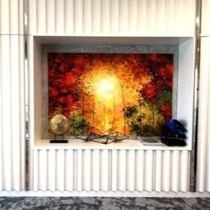 A framed artwork of a sunset with autumn trees, placed on a white shelf with decorative objects on either side, against a beige wall.