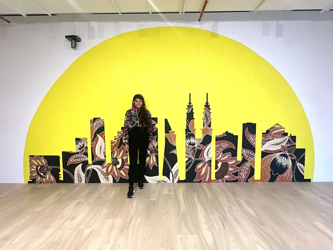 A woman standing in front of a wall mural of a city skyline with a yellow semi-circle background and floral patterns at the bottom.