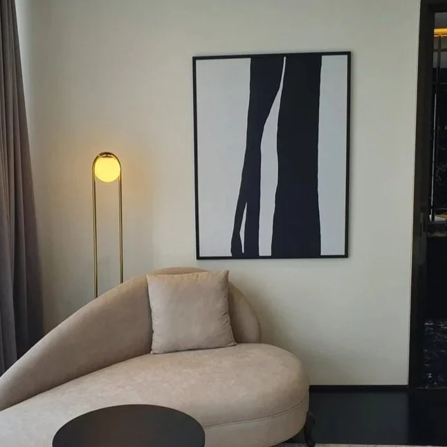 Interior of a modern living room with a beige curved sofa, a matching cushion, a black side table, a tall yellow floor lamp, a framed black and white abstract artwork on the wall, and dark curtains.