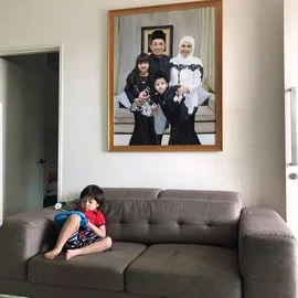 A young girl sitting on a gray sofa in a living room, with a framed family photo hanging on the wall behind her.