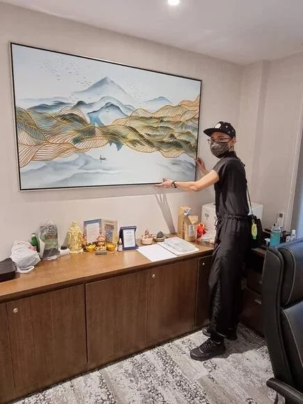 Person wearing a mask and cap, standing next to a large landscape painting of mountains and water, pointing at the artwork in an office or lounge area.