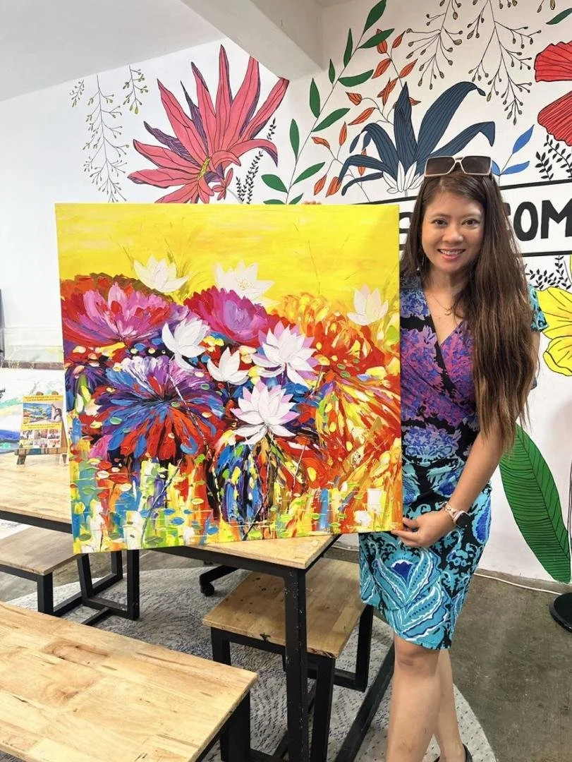 A woman with long brown hair wearing sunglasses on her head and a patterned blue dress is posing next to a colorful abstract floral painting, holding the bottom right corner of the artwork, inside an art gallery with a mural of flowers on the wall.