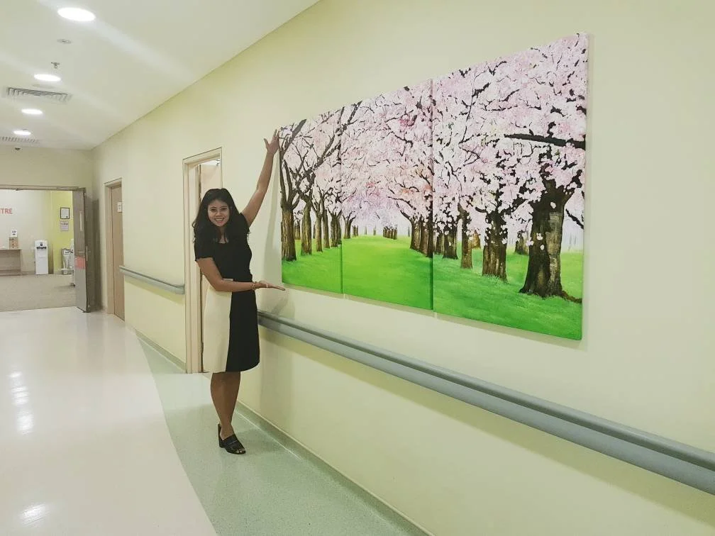 A woman in a black dress and high heels standing in a hallway, smiling, pointing up at a large painting of cherry blossom trees on the wall.