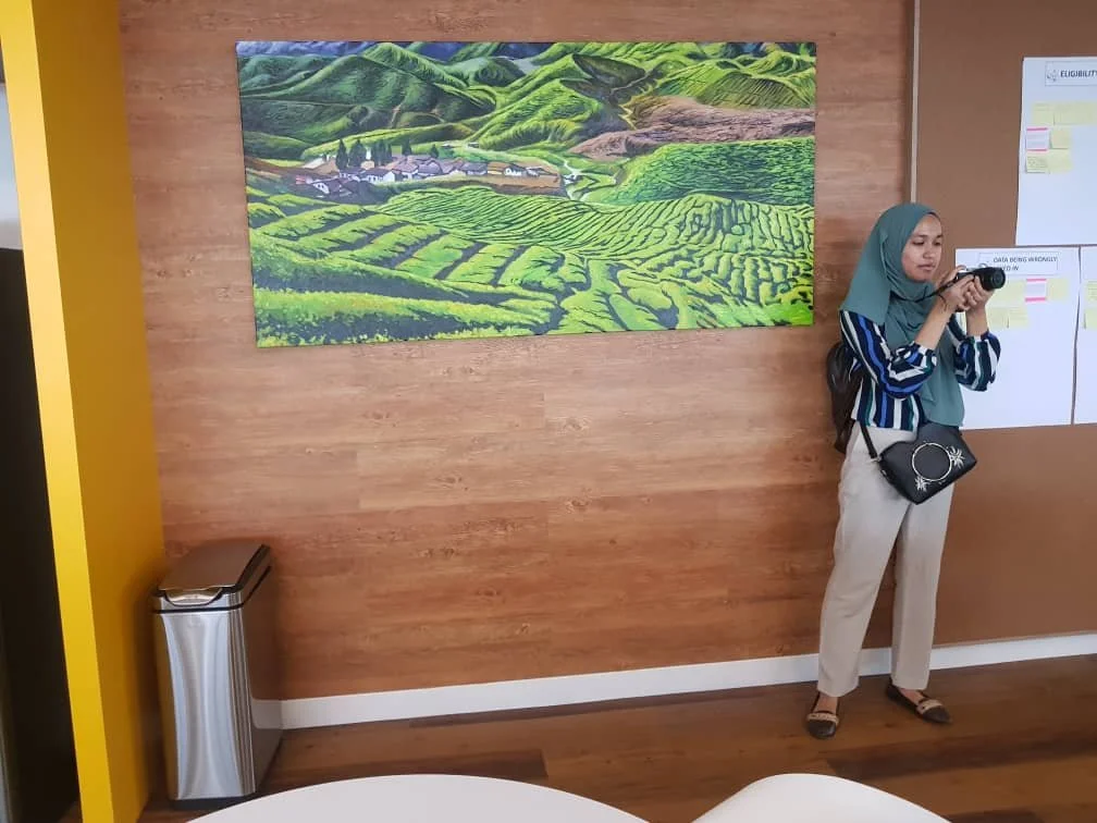 Woman in striped shirt, beige trousers, teal headscarf, and carrying a black bag, taking a photo with a camera inside a room with wooden wall, large landscape painting of green hills and fields, and whiteboards with sticky notes.