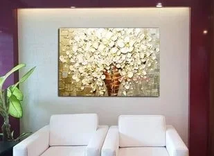 A large framed artwork of white flowers and gold accents hangs above a white sofa in a modern living room. A green potted plant is on the left side of the sofa.