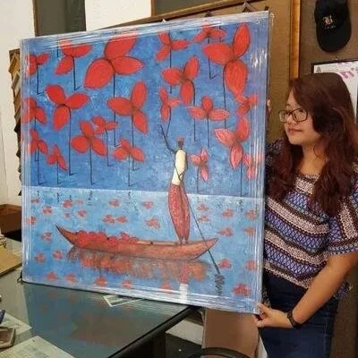 A woman holding a painting of a person in a red boat surrounded by red leaves against a blue sky.