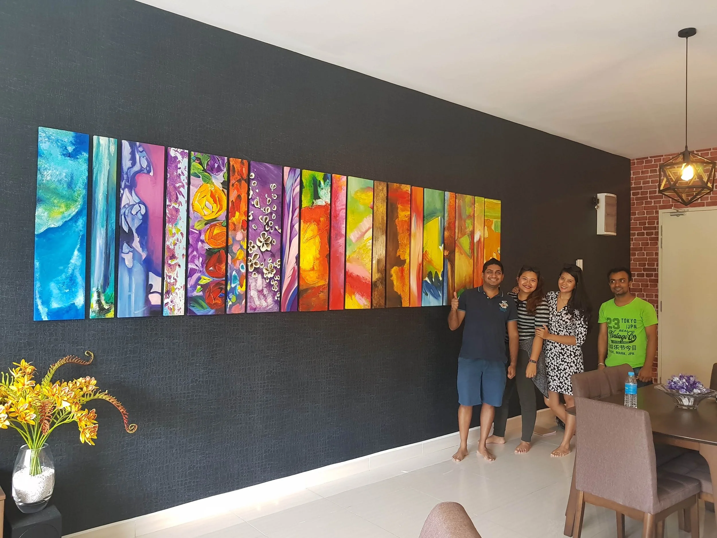 A group of four people standing in front of a colorful abstract art display on a black wall. There are flowers on the table nearby, and the setting appears to be an indoor art gallery or exhibition space.