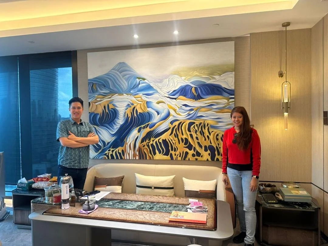 Two people standing in a modern hotel room in front of a large abstract mountain landscape painting. One man wearing a patterned shirt, one woman in a red sweater and jeans. There is a table with various items and a sofa with pillows.