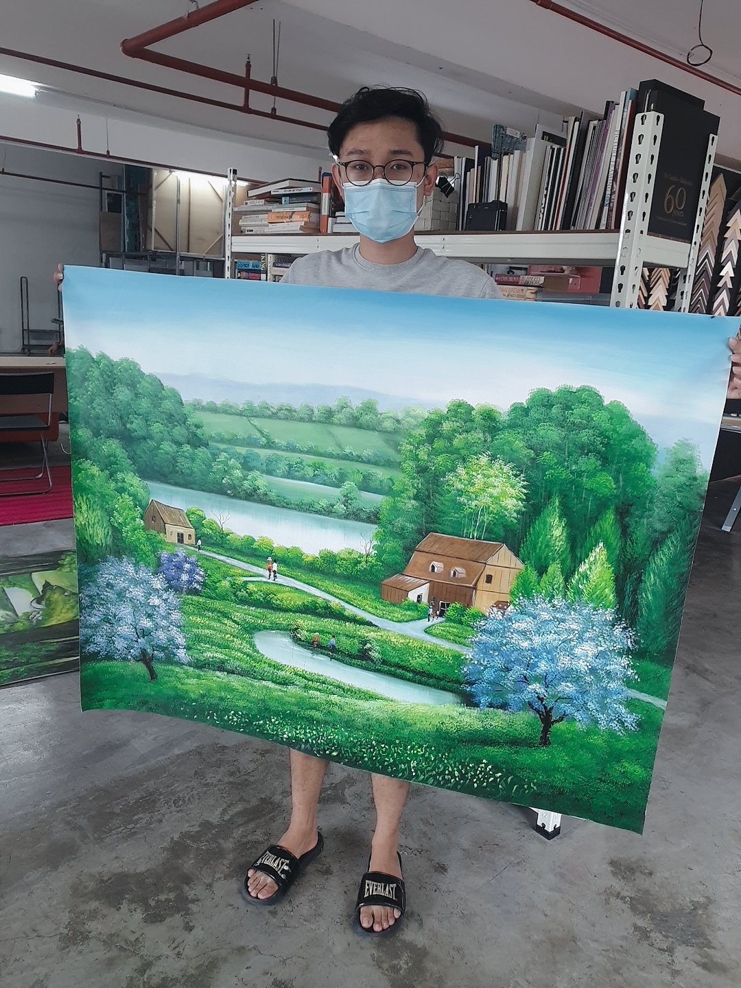 A person wearing glasses and a blue face mask holds a large landscape painting with green trees, a river, and houses, in an indoor space with shelves and books in the background.