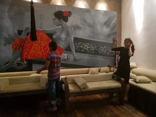 Two children, a boy and a girl, stand in front of a large black-and-white mural of a woman with a red flower in her hair sitting at a table. The children are near a beige sectional sofa in a room with wooden flooring.