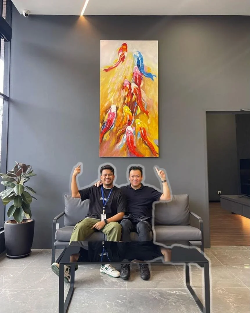 Two men sitting on a gray couch in a modern lobby area, smiling and raising their arms. Behind them is a large, colorful painting of koi fish swimming. To the left is a potted plant, and the floor is tiled.