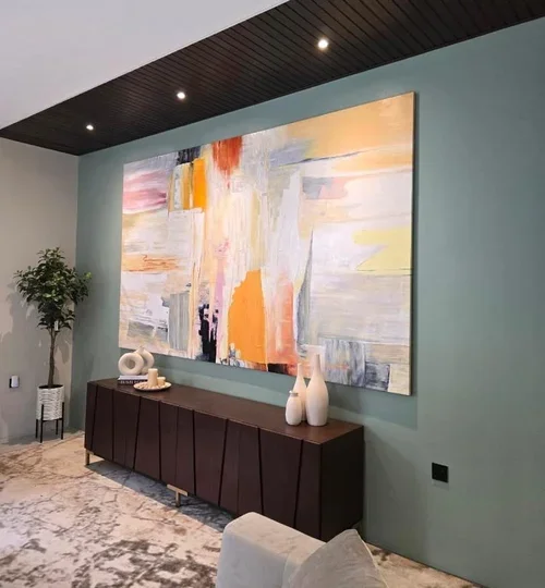 Modern living room with a large abstract painting in orange, yellow, white, and gray tones, a dark wooden sideboard, decorative vases, and a potted plant on a side table.