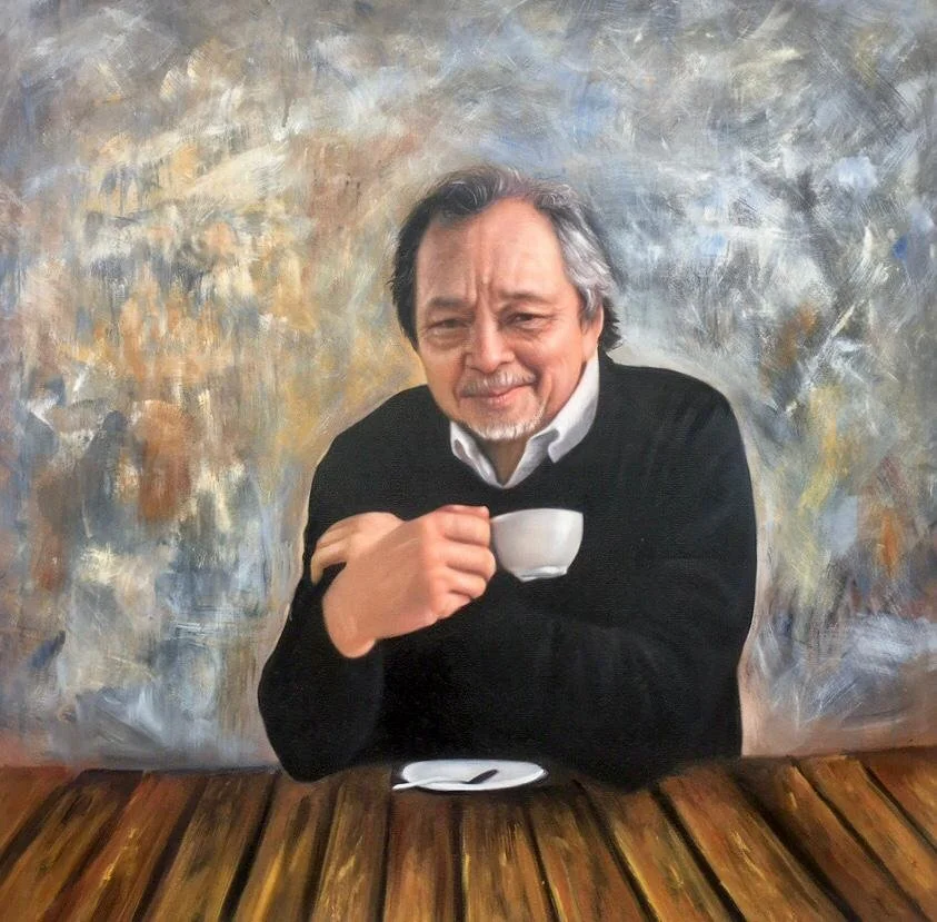 A man with gray hair and a beard, wearing a white shirt and black sweater, sitting at a wooden table with a cup and spoon, smiling at the camera against a painted abstract background.