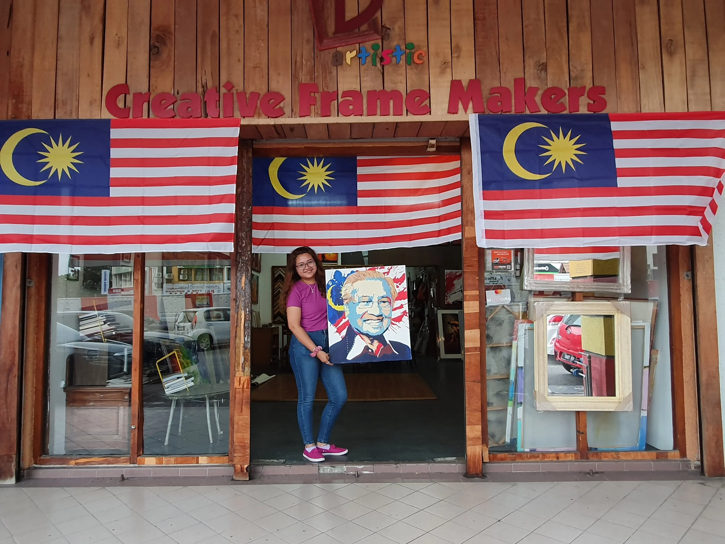 A woman with long hair, glasses, wearing a purple shirt and jeans, is standing inside a wooden art studio, holding a colorful portrait of a man with glasses. The studio is decorated with Malaysian flags and a sign that reads 'Creative Frame Makers.'