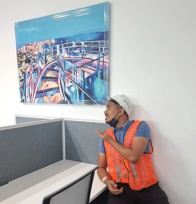 A man in an orange safety vest and white hard hat sitting at a desk and pointing to a colorful abstract painting of a bridge and cityscape on the wall.