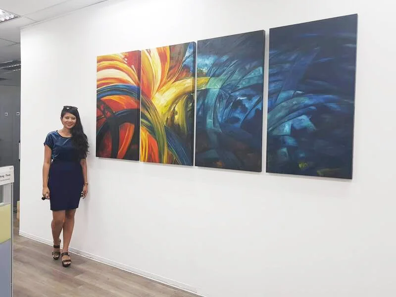 A woman in a navy dress and black heels standing next to a large abstract multi-panel painting with swirling colors, including red, yellow, blue, and green, in an art gallery or exhibit space.