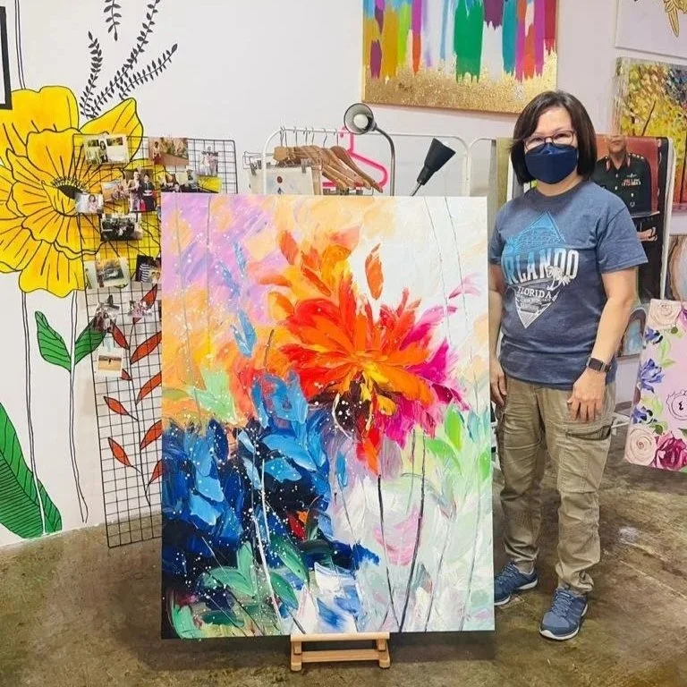 Woman wearing a face mask and glasses standing next to an abstract floral painting. The painting features vibrant colors including orange, red, blue, pink, and green. The background includes various other colorful artwork and decor in an art studio.