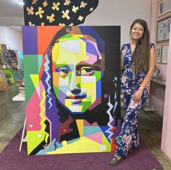 A woman with long hair wearing a blue floral jumpsuit standing next to a colorful, geometric portrait of Mona Lisa on an easel in an art gallery or studio.