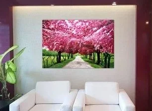 A large wall art print of a tree-lined pathway with blooming pink cherry blossom trees, mounted on a white wall in a living room with two white sofas and a potted plant nearby.