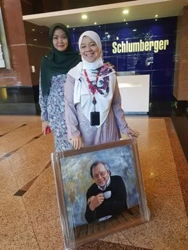 Two women standing in front of a Schlumberger sign, with a framed photo of a man on the floor in front of them.