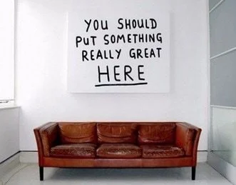 White wall with handwritten sign saying "You should put something really great here" positioned above a brown leather couch in a minimal room.