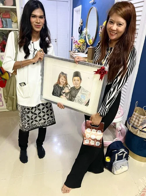 Two women are standing indoors, holding a framed family portrait with a red bow. One woman is wearing a white top and patterned skirt, the other is in a striped black and white top. They are smiling and appear happy.