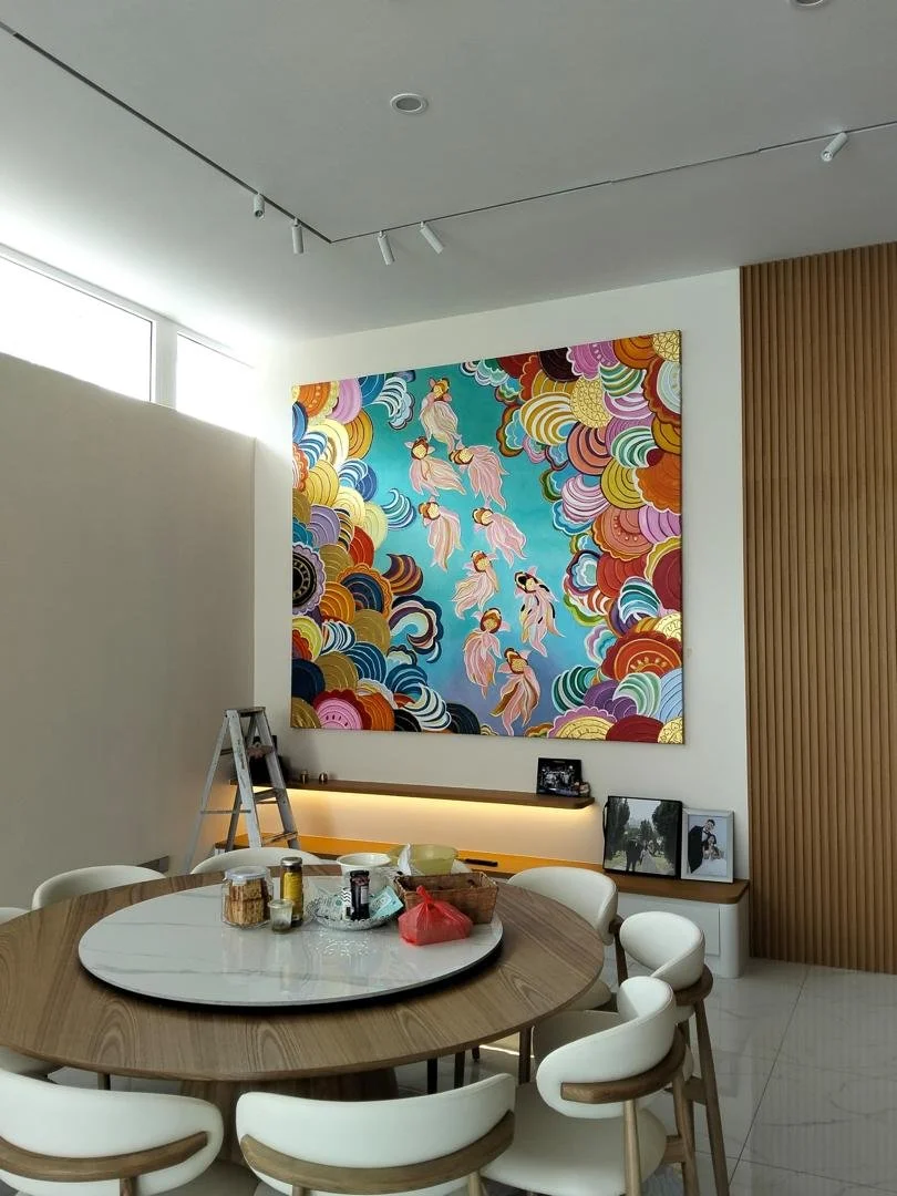 A modern dining room with a round wooden table surrounded by six white chairs, a ladder, some food supplies, and a large colorful artwork of flowing clouds and hanging lanterns on the wall.