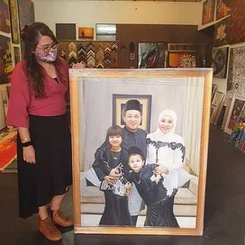 A woman wearing a pink top, black skirt, and face mask standing next to a large framed family portrait of four children and a young man.