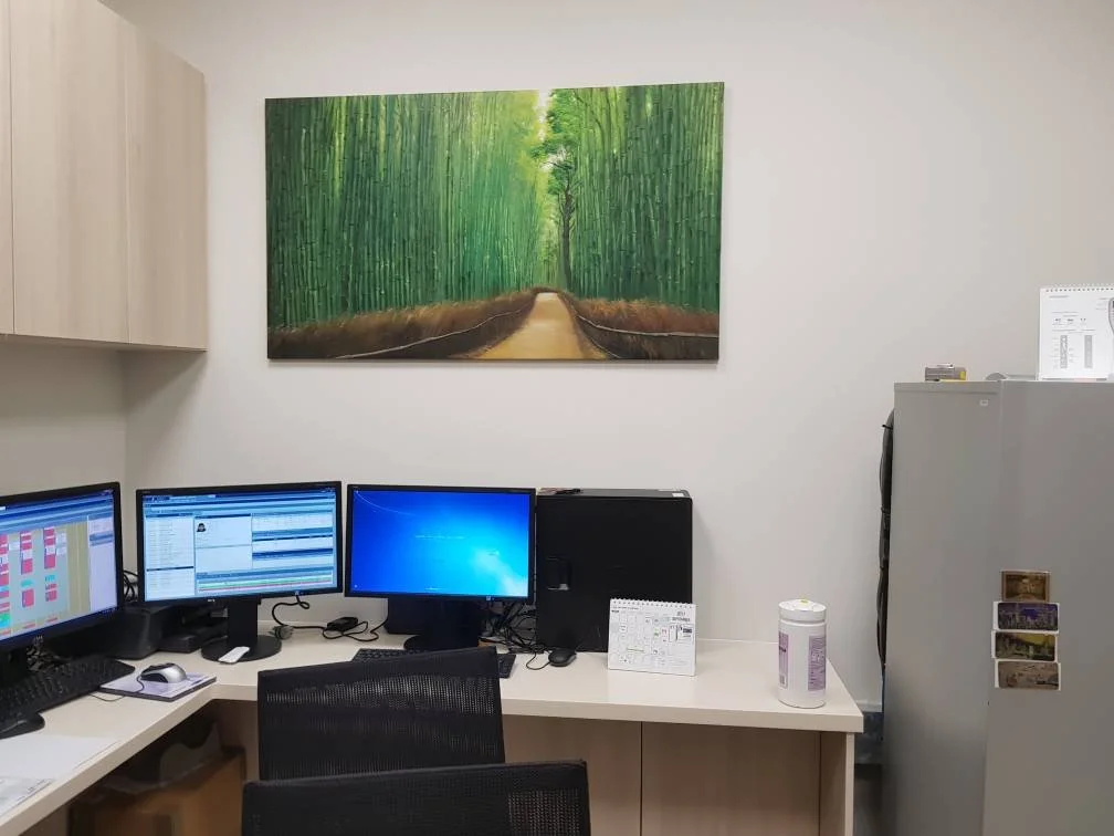 Office workspace with three computer monitors, a black chair, a wooden cabinet on the left, a white wall with a large green forest painting, and a gray storage cabinet with postcards on the right.