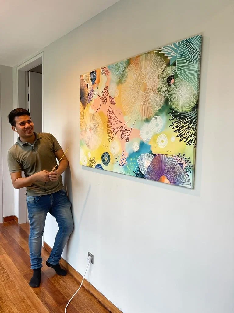 A young man smiling and holding a paintbrush or stick, standing in front of a colorful, abstract floral painting on a wall in a room with hardwood floors.