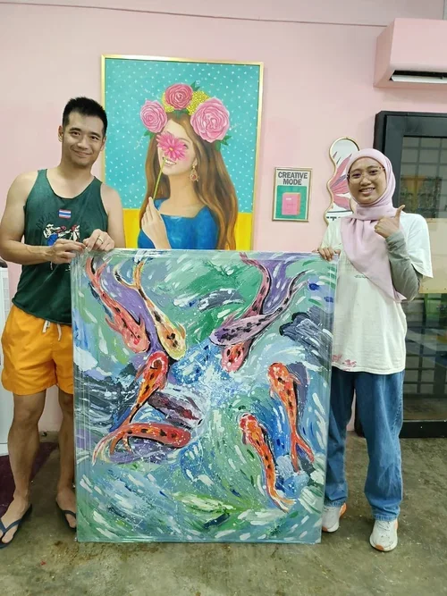 Two people holding a colorful painting of fish swimming in water, standing in art studio with bright artwork and wall decorations.