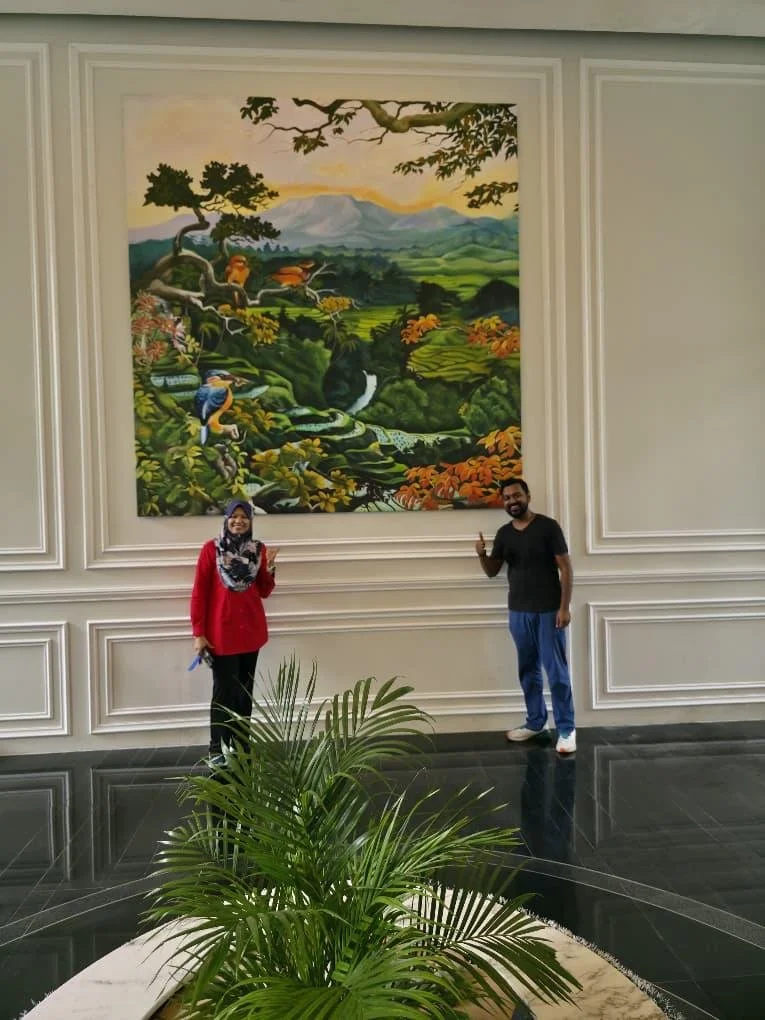 Two people pose in front of a large landscape painting at an art gallery, with a plant in the foreground.
