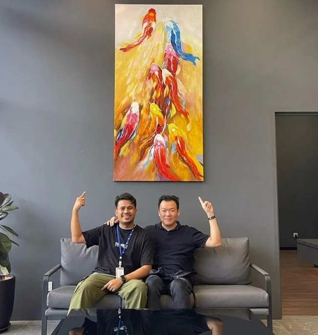 Two men sitting on a gray couch in front of a gray wall, smiling and pointing upward. A colorful abstract painting of parrots hangs on the wall behind them.
