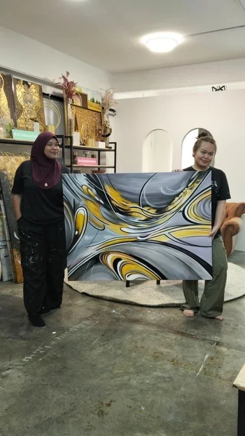 Two women holding an abstract painting with swirls of gold, gray, black, and white inside a room decorated with gold accents and art pieces.