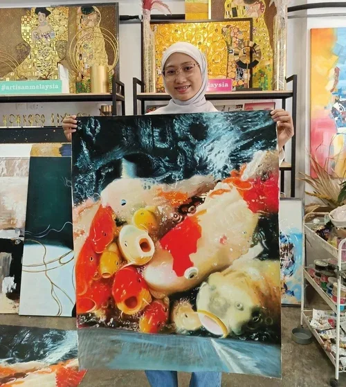 A woman wearing a white hijab and glasses is holding a colorful painting of koi fish swimming in water. She is smiling and standing in an art studio with various artworks and supplies in the background.
