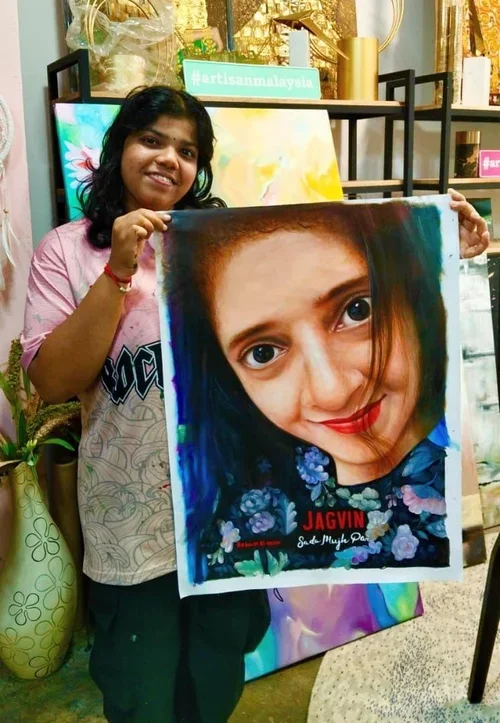 A woman with dark hair and a pink shirt is smiling and holding a large colorful portrait of a woman's face with brown eyes and red lipstick. The background includes art supplies and decorated items.