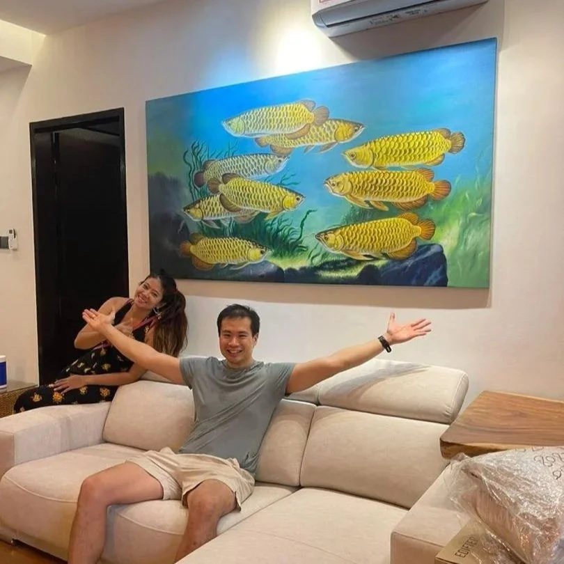 Two people sitting and smiling on a beige sofa in a living room, with a colorful painting of fish and underwater plants hanging on the wall above them.