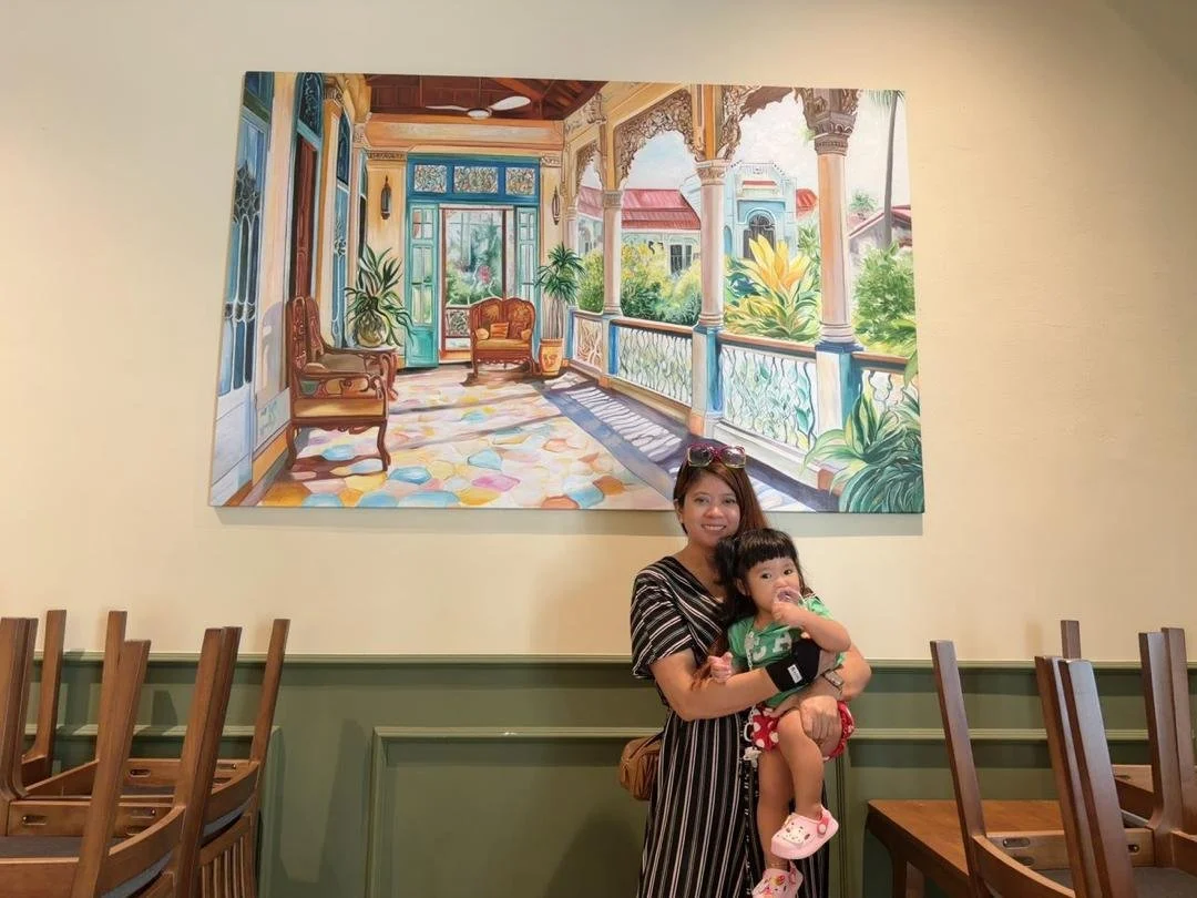 A woman holding a young girl in a restaurant with a large colorful painting of a porch or veranda hanging on the wall behind them.