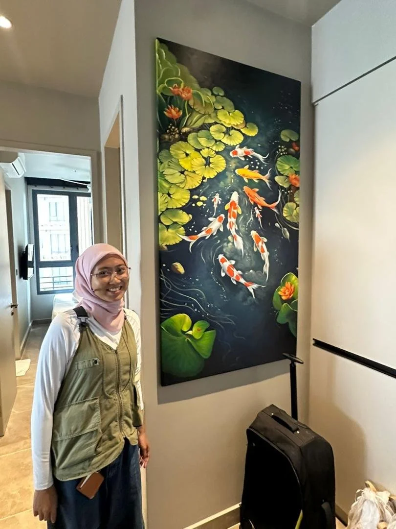 A woman with glasses wearing a pink headscarf, white long sleeve shirt, and green vest standing next to a large painting of koi fish swimming among lily pads and flowers in water.
