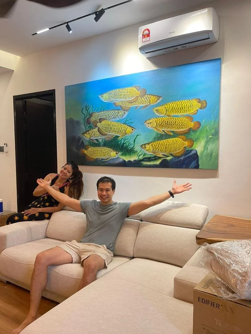 Two people sitting on a beige couch in a living room with a large fish painting on the wall behind them. The man is sitting with arms open wide, and the woman is smiling and leaning back. An air conditioner is mounted above the painting.