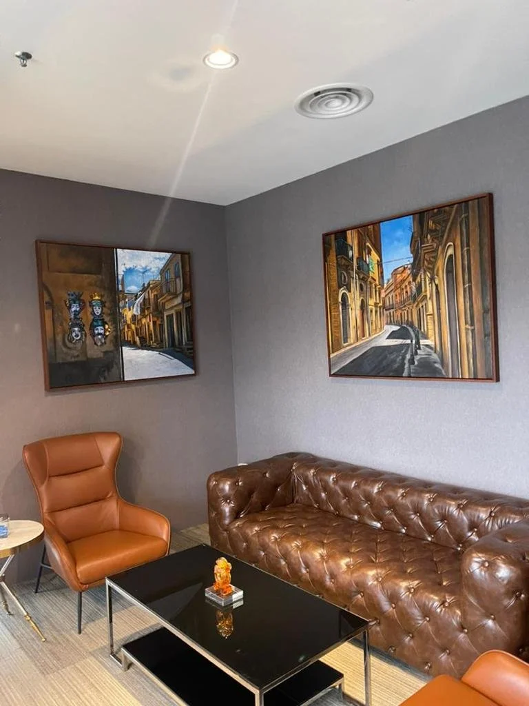 Living room corner with a brown leather tufted sofa, a brown armchair with a high back, a black coffee table, and two framed paintings of city streets hanging on gray walls.