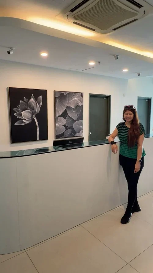 A woman stands in a modern, well-lit lobby or reception area, leaning on a white reception desk. Behind her are two black-and-white floral artwork pieces on the wall, and a door is visible next to the artworks. The ceiling has recessed lighting and a