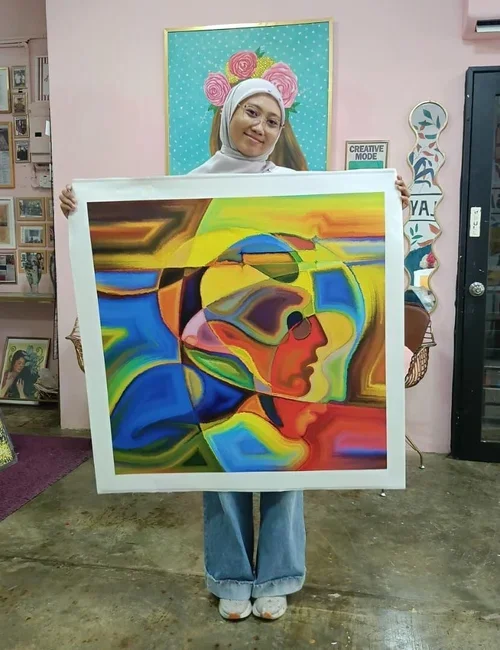 A woman standing indoors, holding a colorful abstract portrait painting of a person wearing a cap, with a background of pink, yellow, blue, and green hues. She is wearing a white headscarf, glasses, a white top, and blue jeans.