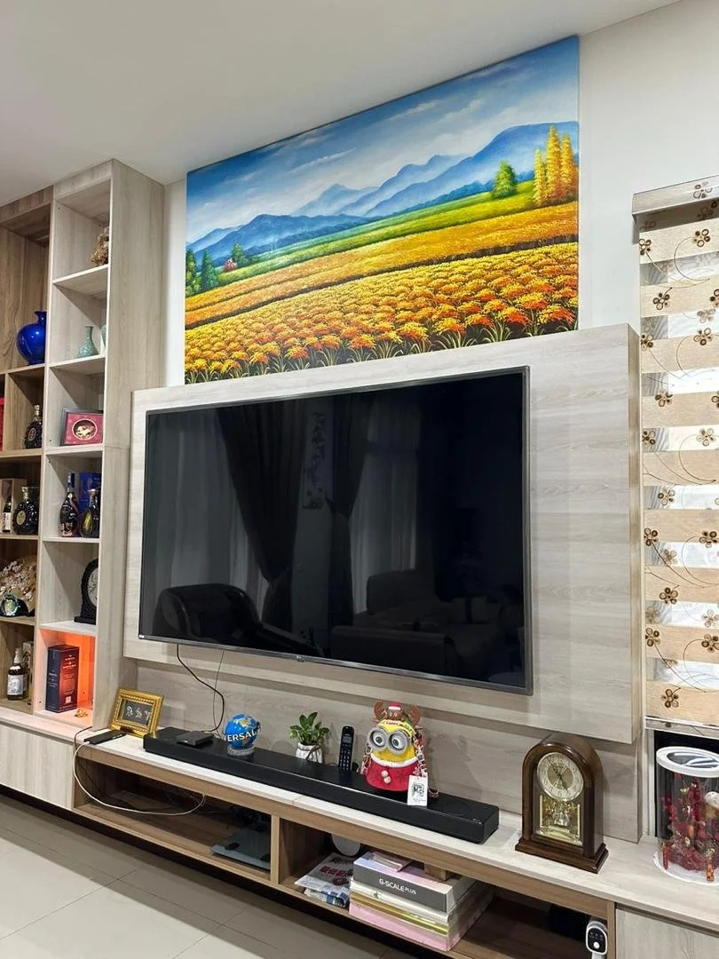 A living room with a large flat-screen TV mounted on a light-colored wooden wall panel. Above the TV, there is a colorful landscape painting of mountains, green hills, and a field of yellow and orange flowers. To the left, there is a tall bookshelf f