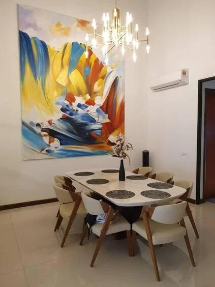 A modern dining room with a rectangular table surrounded by six chairs. The table has a black vase with dried flowers as a centerpiece. There is a large, colorful abstract painting on the wall behind the table and a contemporary chandelier hanging ab
